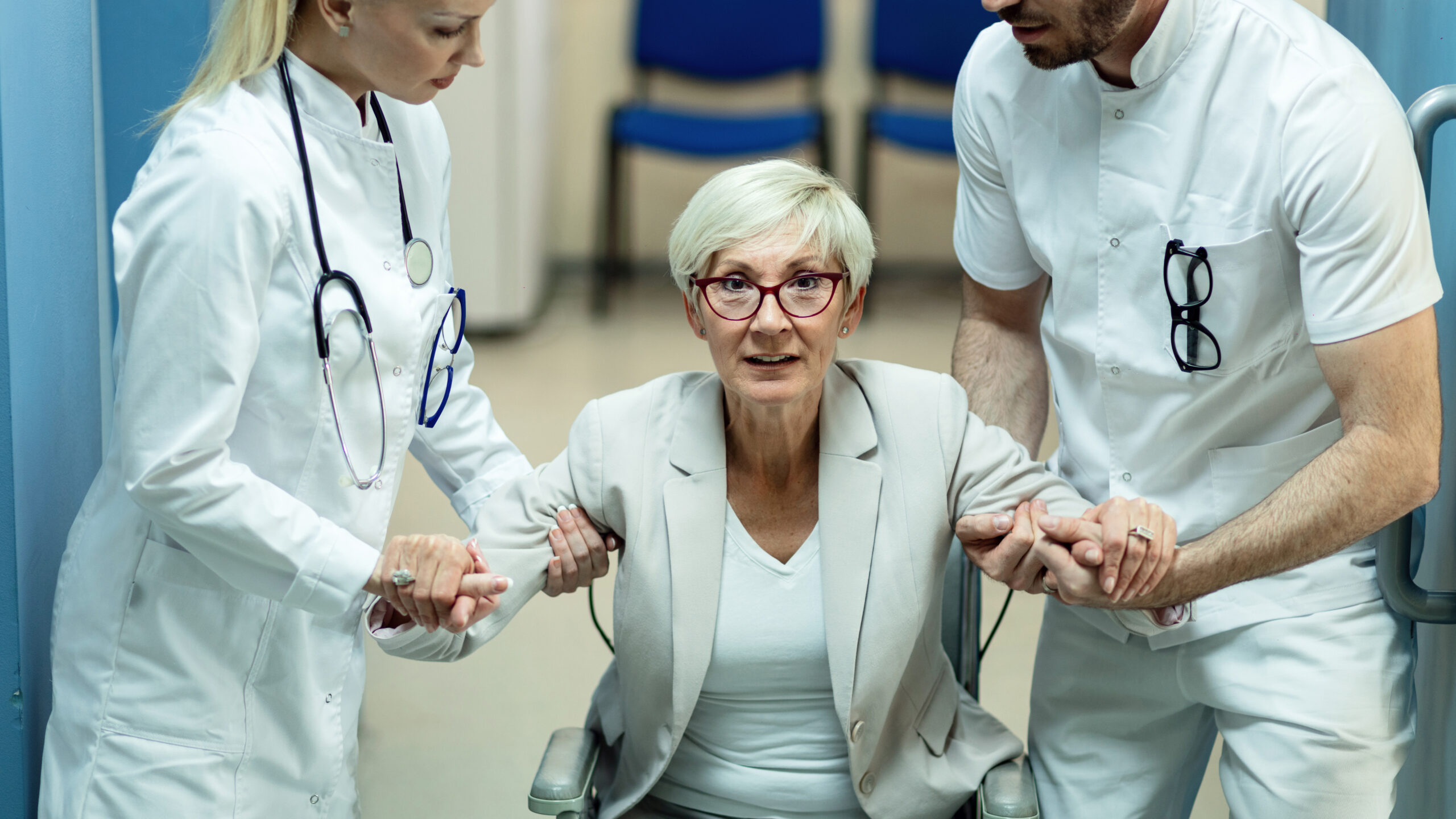 Disabled mature woman getting up from the wheelchair with help of two doctors at clinic.