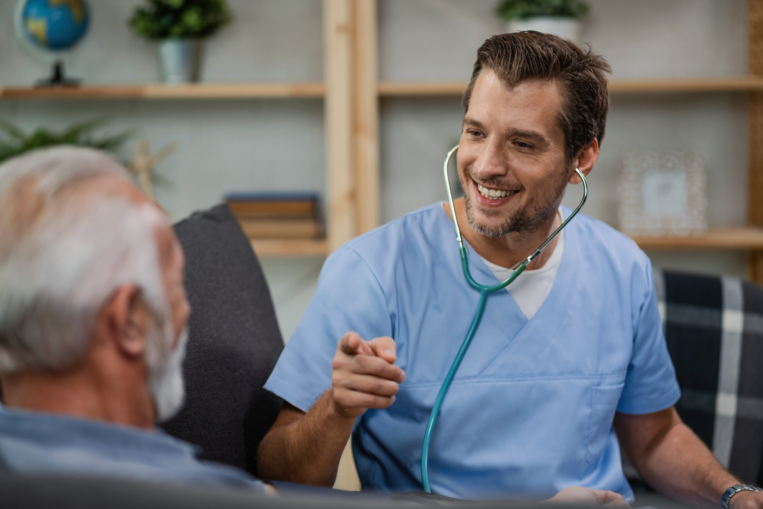 Happy healthcare worker communicating with senior man and pointing at him while being in a home visit.
